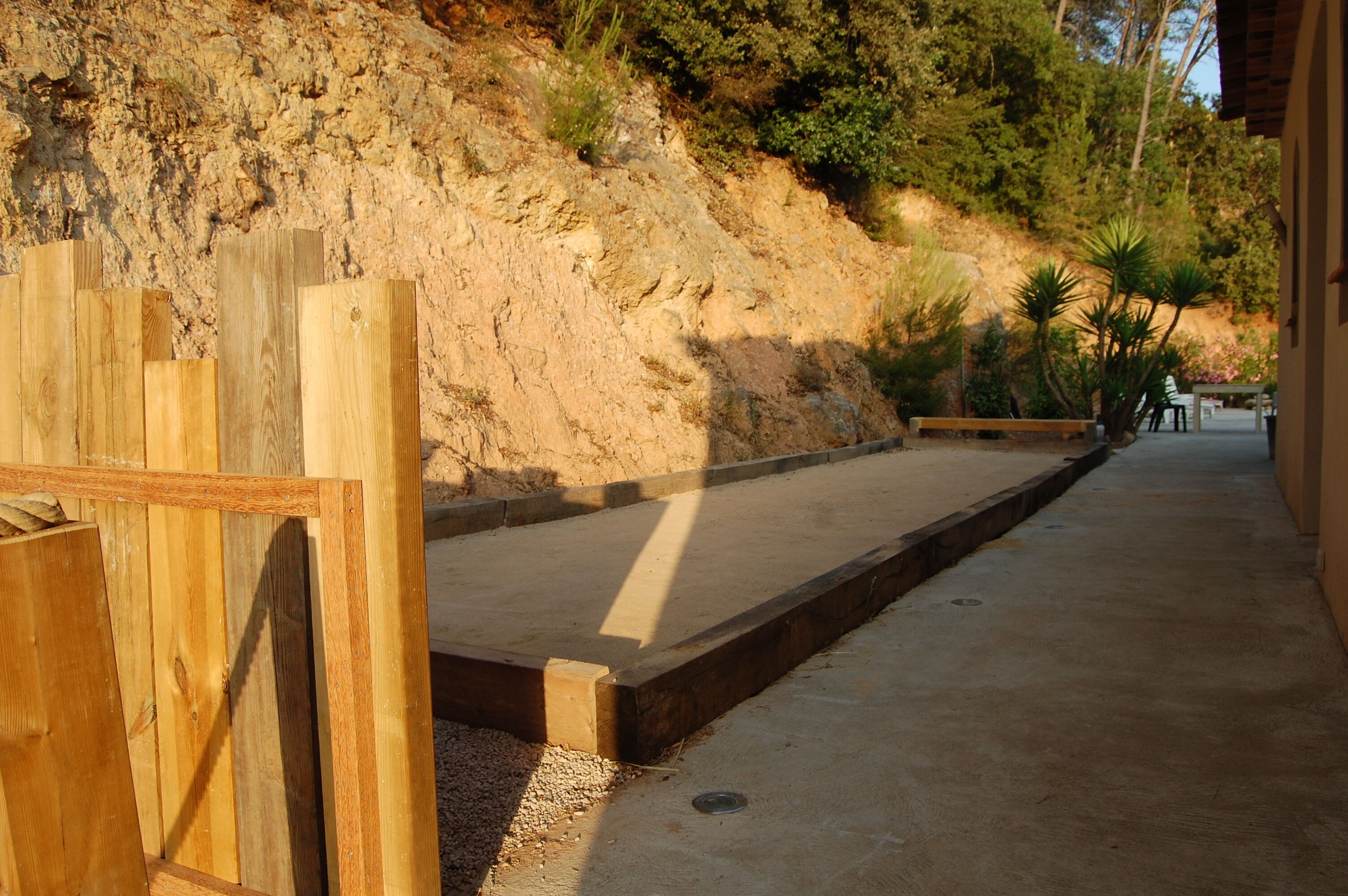 Un sentier mène à un espace aménagé avec des sièges, bordé d’une clôture en bois d’un côté et avec une colline rocheuse en arrière-plan.