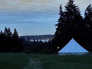 A tent in a grassy field with a view of a distant cityscape and a moon in the sky.