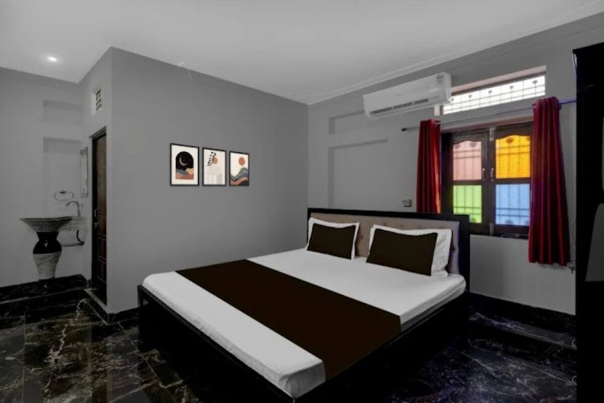 Une chambre à coucher avec un lit, une fenêtre avec des rideaux, un climatiseur fixé au mur et une petite table sur laquelle se trouve un vase.