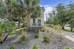 A two-story house with a balcony, surrounded by a gravel garden with various plants and a palm tree.