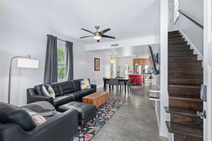 A modern living room with a black leather sofa, a wooden coffee table, and a patterned rug. A dining area with a table and chairs is visible in the background. A staircase with wooden steps leads to an upper level. - Lincolnville Charm l Walk to DNTN l Dog Friendly (St. Augustine)