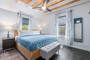 A bedroom with a wooden bed, a bench, a nightstand, a ceiling fan, and two windows with curtains. - Lincolnville Charm l Walk to DNTN l Dog Friendly (St. Augustine)