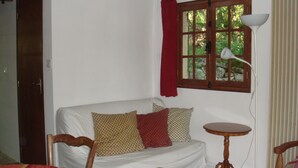 Ett rum med en vit soffa, en trästol, ett litet runt bord, en lampa och ett fönster med en röd gardin.