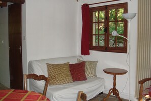 Ein Zimmer mit einem weißen Sofa, einem Holzstuhl, einem kleinen runden Tisch, einer Lampe und einem Fenster mit rotem Vorhang.