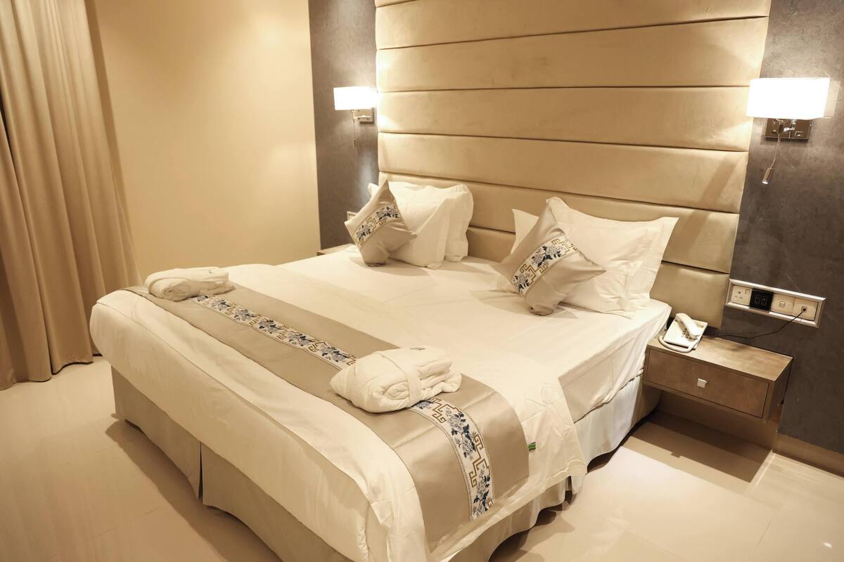 Quarto de hotel com uma cama grande, uma mesinha de cabeceira, uma luminária e uma tomada.
