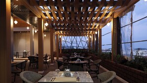 An outdoor dining area with tables and chairs, a wooden pergola, and large windows offering a view. - Al Masa Al Taj Hotel Apartments (Ha'il)