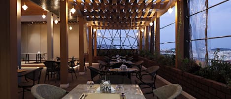 Un espacio de comedor al aire libre con mesas y sillas, una pérgola de madera y amplias ventanas que ofrecen vistas.