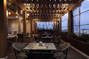 An outdoor dining area with tables and chairs, a wooden pergola, and large windows offering a view.