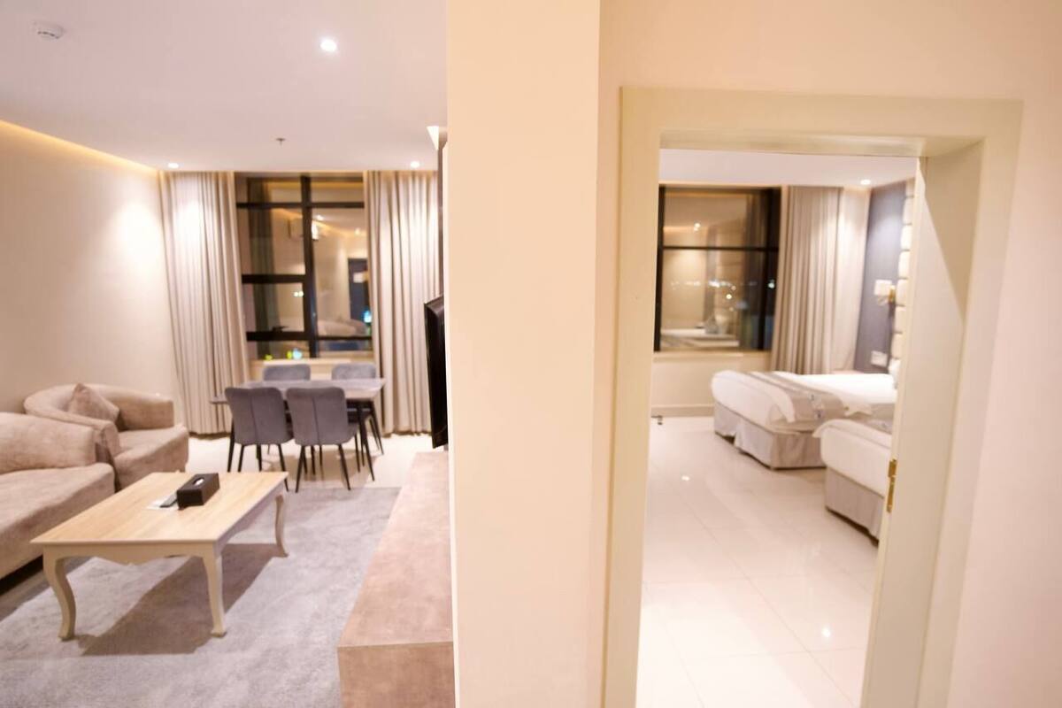 Quarto de hotel com sofá, mesa de jantar com cadeiras, pequena cozinha e quarto separado com cama.