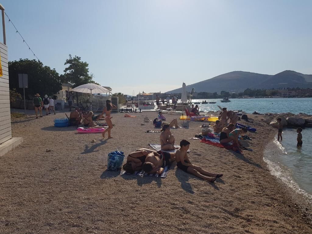 A beach with people sunbathing and swimming, a building on the left, and mountains in the background.