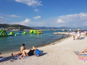 Una platja amb gent prenent el sol i banyant-se, un pont al fons i un cel serè.