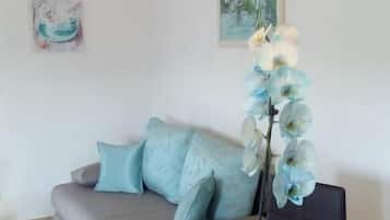 A modern living room with a glass coffee table, a sofa with cushions, a tall plant, and a painting on the wall.