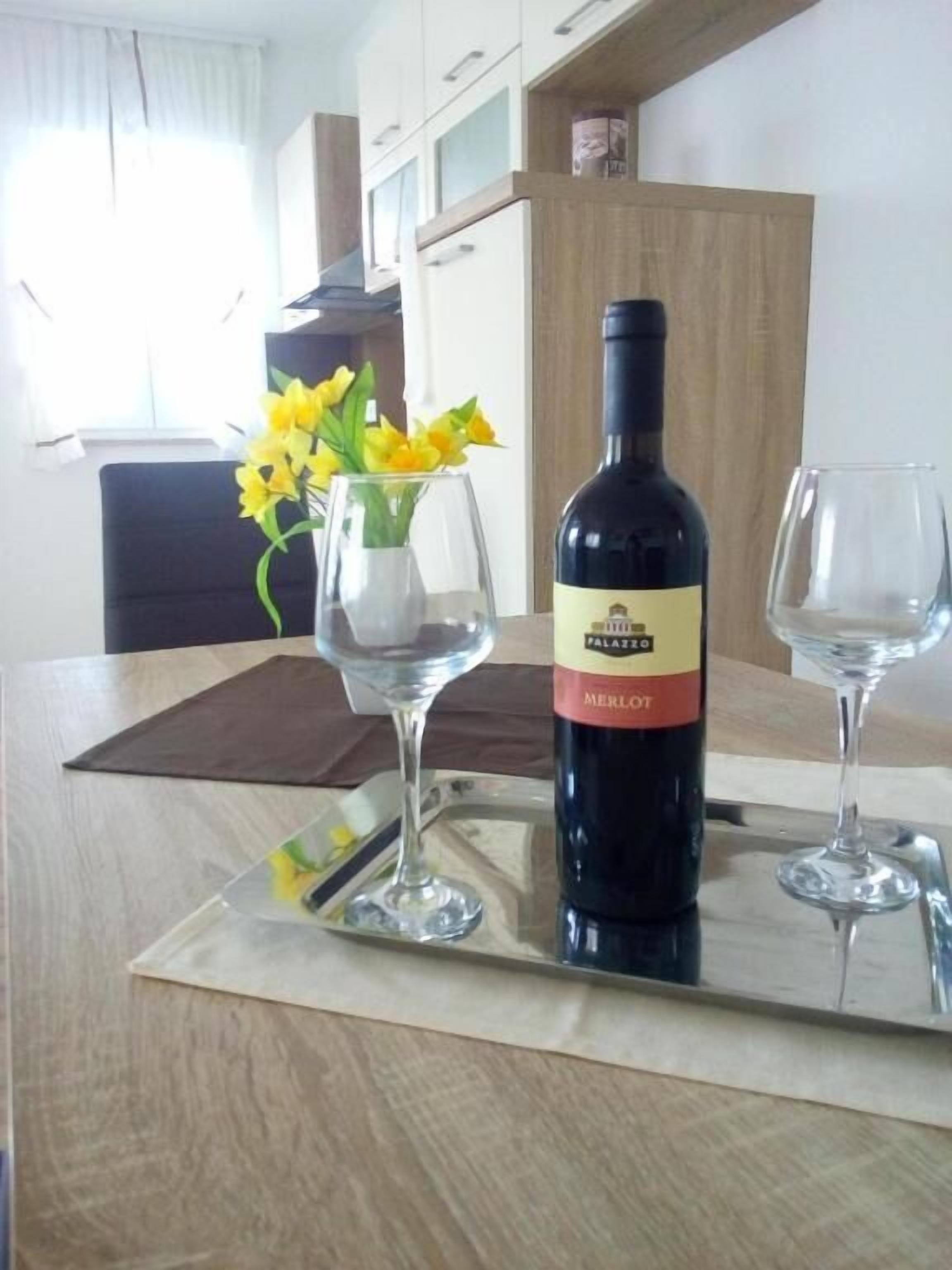 A bottle of wine on a tray with two glasses, a vase with yellow flowers, and a kitchen area in the background.