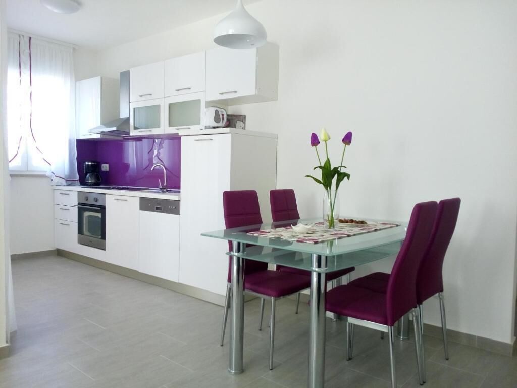 A modern kitchen with white cabinets, a glass dining table, and purple chairs.