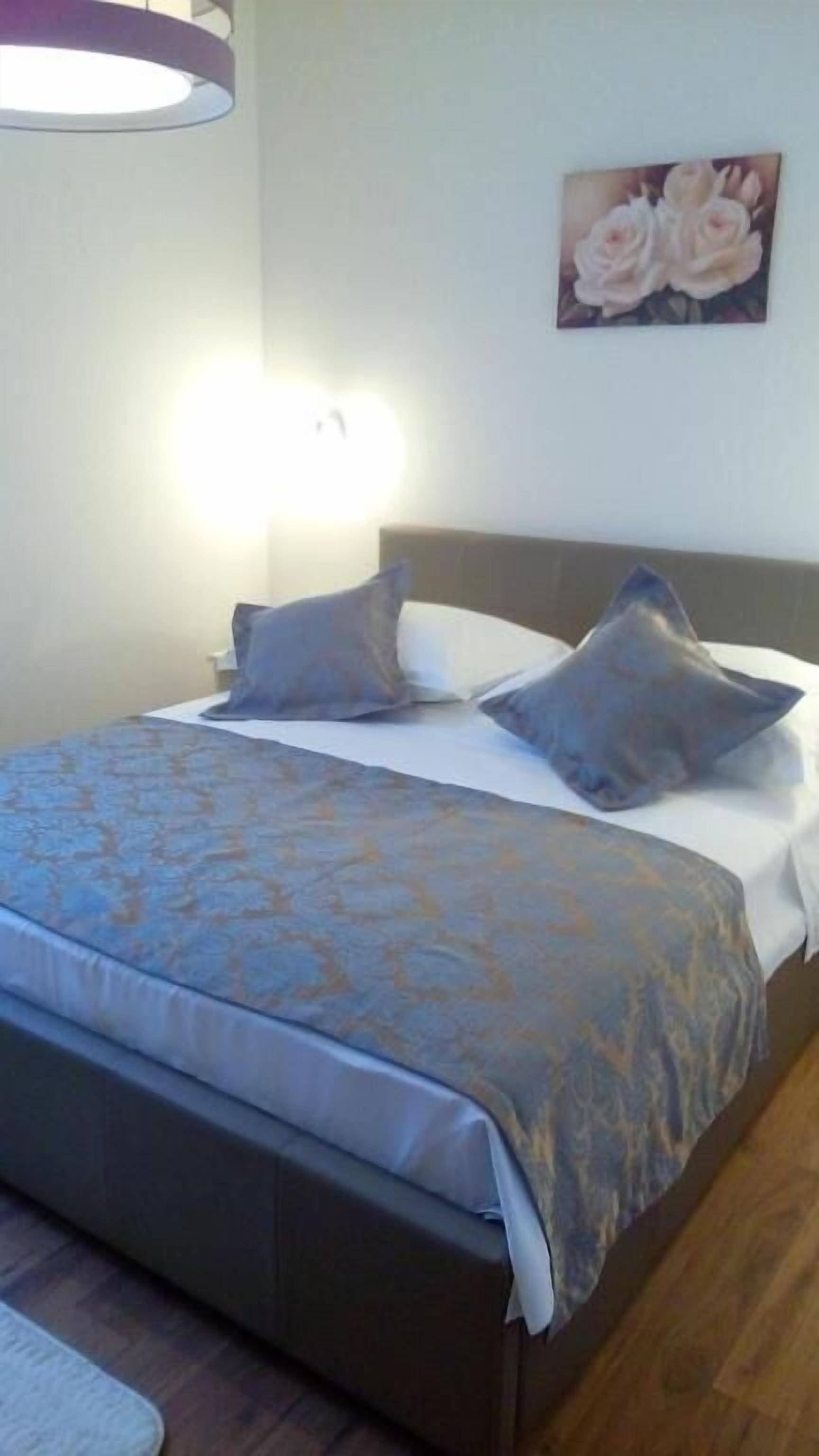 Uma cama com colcha estampada, duas almofadas, uma cabeceira e um candeeiro fixado na parede.