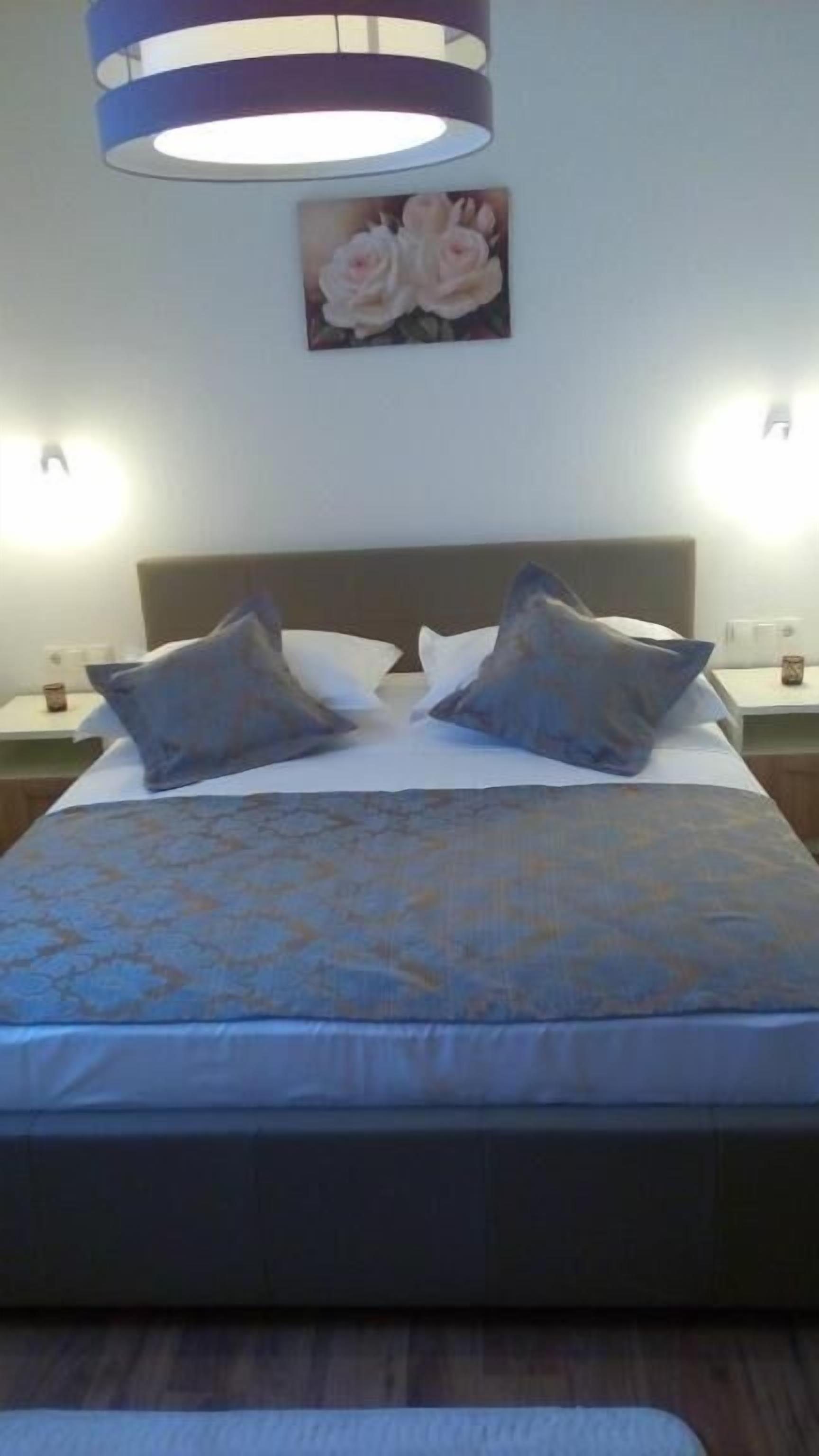 A bed with a patterned bedspread, two pillows, a headboard, and a wall-mounted picture of roses.