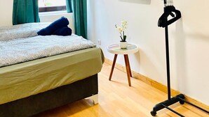 Un dormitorio con cama, ventana con cortinas, una mesita con una planta y una obra de arte en la pared.