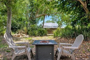 Two Adirondack chairs facing a grill table with a metal grill cover, surrounded by lush greenery and a wooden structure in the background. - The Ashton Hideaway (Sarasota)