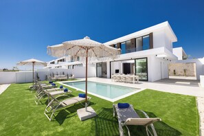 A modern outdoor pool area with lounge chairs and umbrellas, a pool, and a contemporary building with large windows. - Brand New 4 Bedroom Villa with private pool, 100 meters from the beach (Protaras)