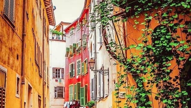 Eine enge Straße mit Gebäuden an beiden Seiten, ein Café mit Außenplätzen und eine sichtbare Treppe, die zu einem Obergeschoss führt.