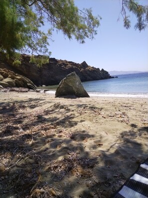 A beach with a large rock in the foreground, clear blue water, and rocky cliffs in the background.