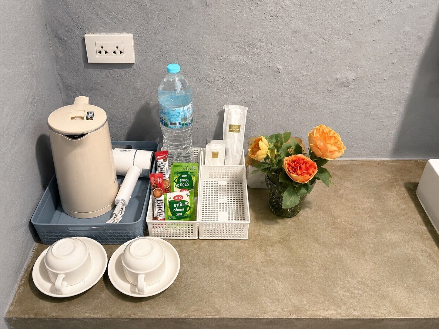 A neatly arranged hotel room with a trash bin, a water bottle, a small vase with flowers, a tissue box, and a basket with toiletries.