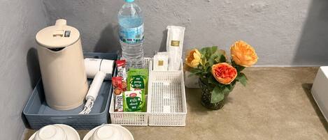 A neatly arranged hotel room with a trash bin, a water bottle, a small vase with flowers, a tissue box, and a basket with toiletries.