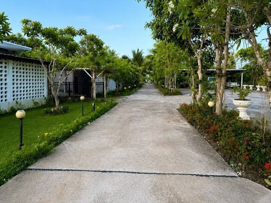 A concrete pathway flanked by greenery and buildings, leading to a structure with a covered entrance.