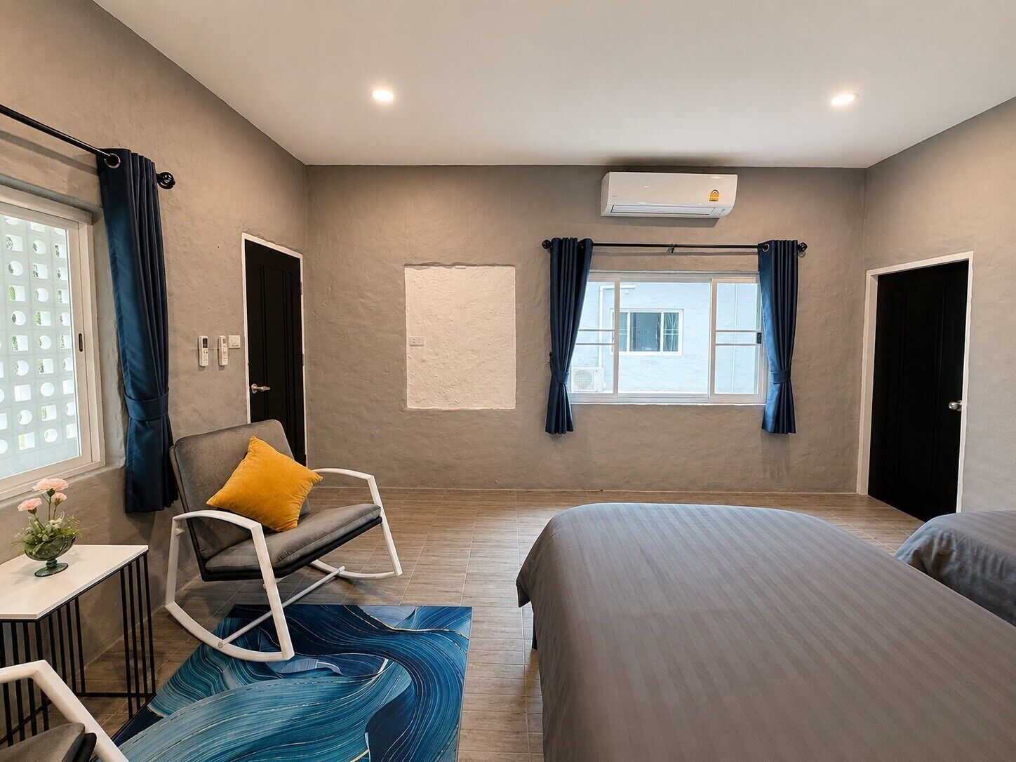 Un dormitorio con cama, una silla, una mesita, ventana con cortinas, aire acondicionado y una puerta.