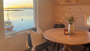 Un intérieur en bois avec une table ronde, un luminaire suspendu et une vue sur un bateau, visible à travers une grande fenêtre.