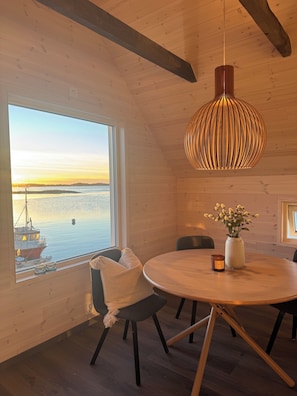 Un intĂ©rieur en bois avec une table ronde, un luminaire suspendu et une vue sur un bateau, visible Ă travers une grande fenĂȘtre.