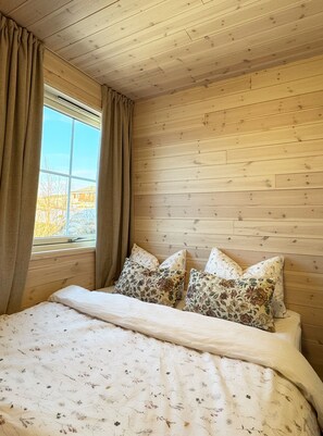 A wooden-paneled room with a bed, floral-patterned pillows, and a window with curtains. - New and exclusive cabins with fantastic sea views, right by the sea. (Nordland)