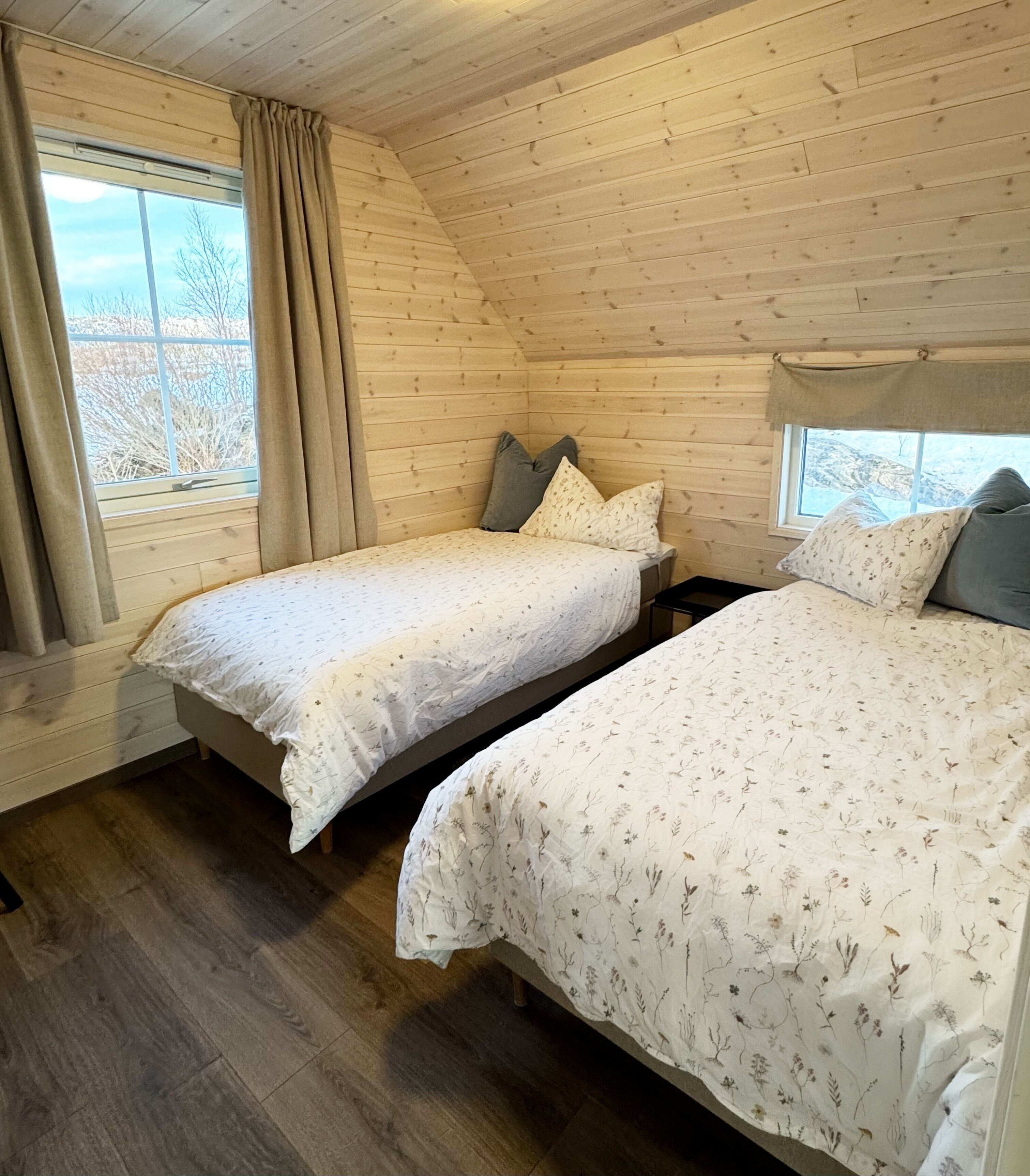 Een kamer met twee bedden, houten wanden, een raam met gordijnen en uitzicht op bomen.