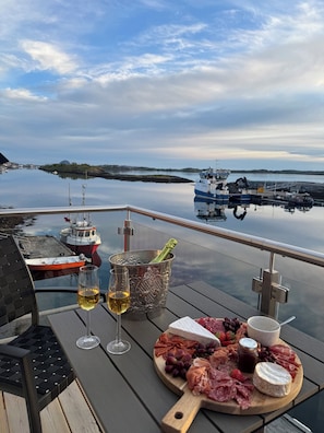 A wooden deck with a glass table, a bottle of champagne, two glasses of wine, a charcuterie board with meats, cheeses, and grapes, and a view of boats on calm water. - New and exclusive cabins with fantastic sea views, right by the sea. (Nordland)