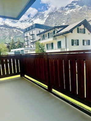 A balcony with a view of snow-capped mountains, a wooden fence, and a building with multiple windows and balconies. - Lovely vacation apartment with sun & mountain view (Saas-Fee)