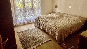 Een slaapkamer met een bed, vloerkleed, stoel en een raam met gordijnen.