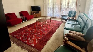Een woonkamer met een roodgekleurd tapijt, een blauwe bank, een rode fauteuil, een houten salontafel en een televisie op een houten dressoir.