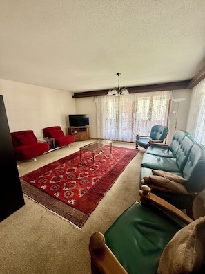 A living room with a red carpet, a blue sofa, a red armchair, a wooden coffee table, and a television on a wooden stand. - Lovely vacation apartment with sun & mountain view (Saas-Fee)
