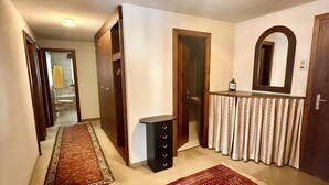 Een gang met een kamerbreed tapijt, een klein zwart kastje, een spiegel en een deur naar een andere kamer.