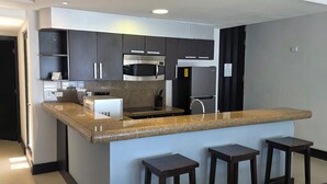 Una cocina moderna con armarios oscuros, encimera de granito, microondas integrado y refrigerador de acero inoxidable. Hay tres taburetes de madera en la barra.