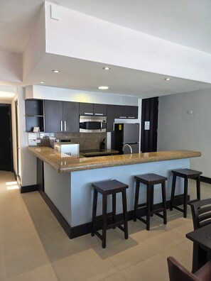 A modern kitchen with dark cabinetry, a granite countertop, a built-in microwave, and a stainless steel refrigerator. There are three wooden stools at the counter. - Aldea Thai, Ocean front penthouse w/ private rooftop patio & pool w/ ocean view. (Playa del Carmen)