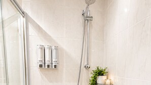 Un área de ducha con puerta de vidrio, un cabezal de ducha de mano, un grifo empotrado en la pared y una repisa con artículos de aseo y una planta.