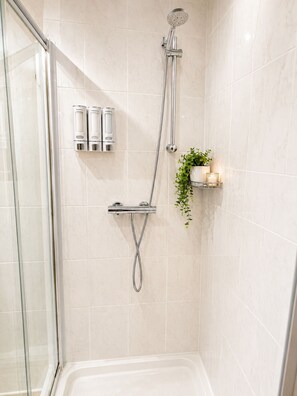 A shower area with a glass door, a handheld showerhead, a wall-mounted faucet, and a shelf with toiletries and a plant. - 
   Countryside Studio Retreat on 13 Acres of Private Farmland (Chelmsford)