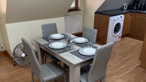 Un comedor con una mesa dispuesta para cuatro, sillas grises, un ventilador y una lavadora al fondo.