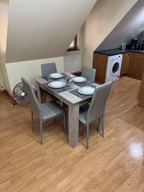 Un coin repas avec une table dressée pour quatre personnes, des chaises grises, un ventilateur et, au fond, un lave-linge.