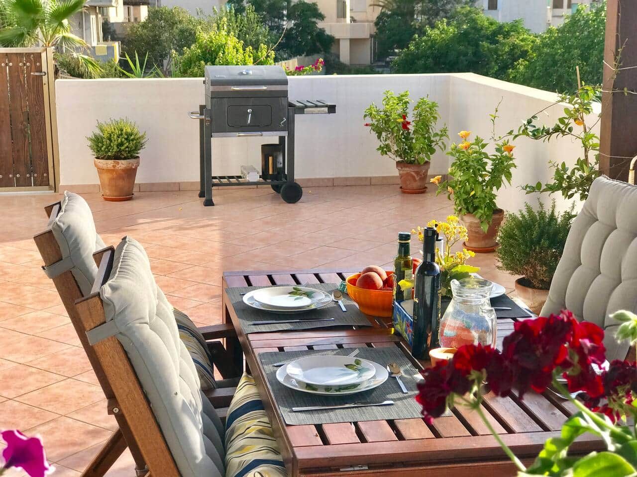 En udendørs terrasse med spiseplads til to, grill og plantekasser.