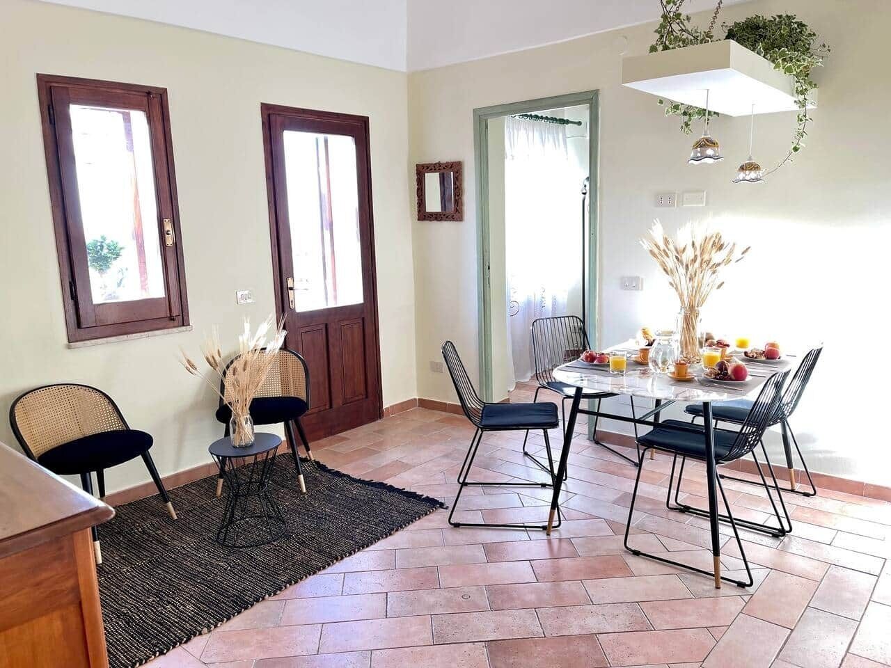 Un comedor con una mesa dispuesta para cuatro personas, rodeada de sillas, una alfombra y una puerta que da a otra habitación.