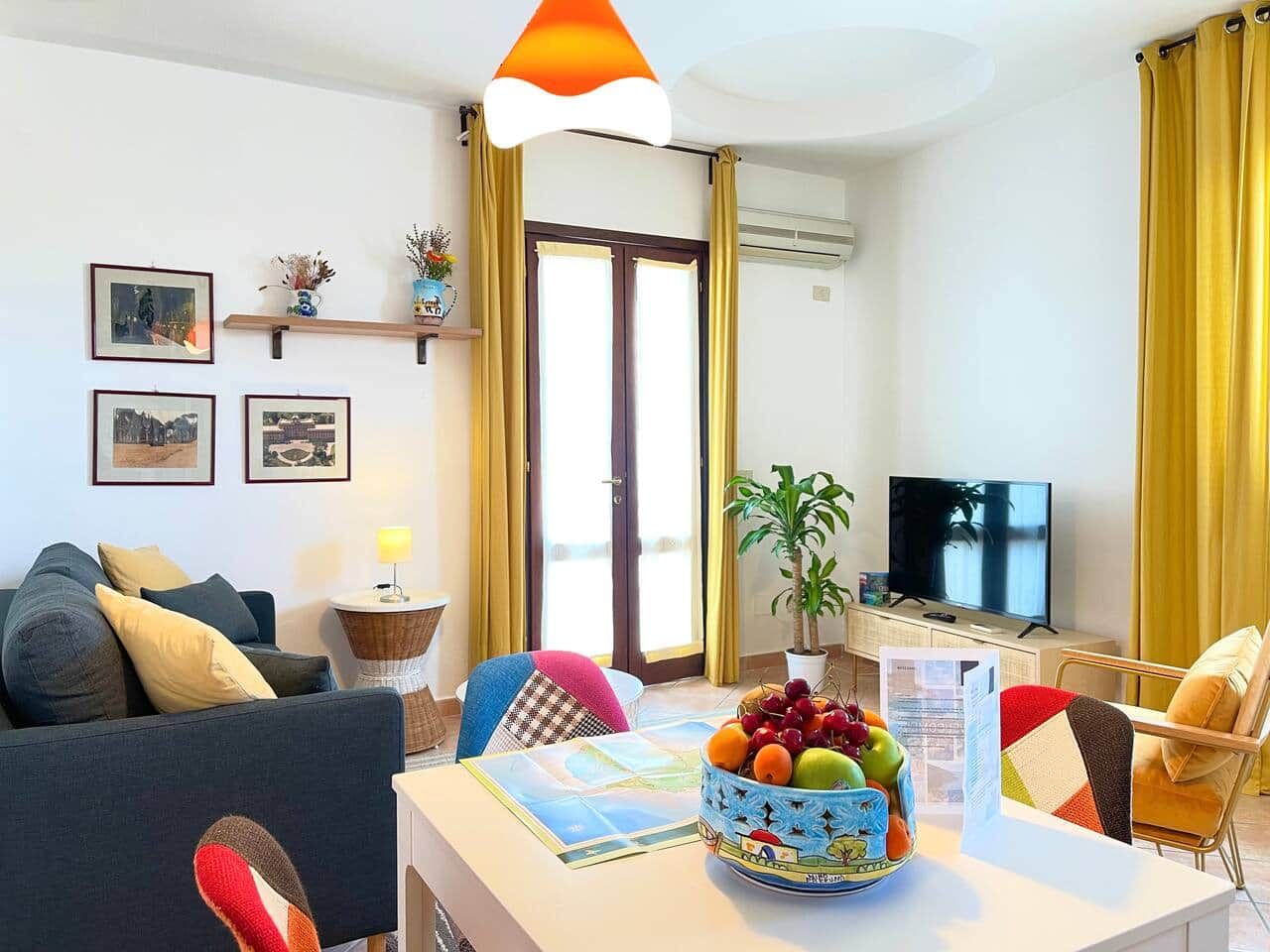 A living room with a sofa, a dining table with a fruit bowl, a television, and a plant.