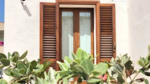 A building with a window featuring wooden shutters, a security camera, and some greenery in the foreground.