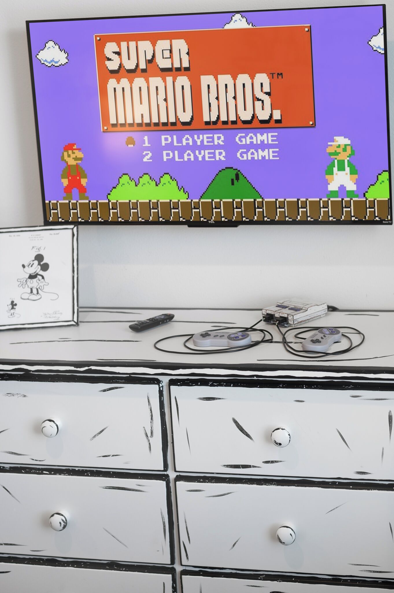 Et Super Mario Bros. spil på skærmen, et indrammet Mickey Mouse billede og en hvid kommode med skuffer.